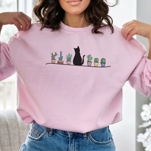 May include: A pink sweatshirt with a black cat sitting in front of a row of potted cacti. The cat is facing left and the cacti are in brown pots with white accents.