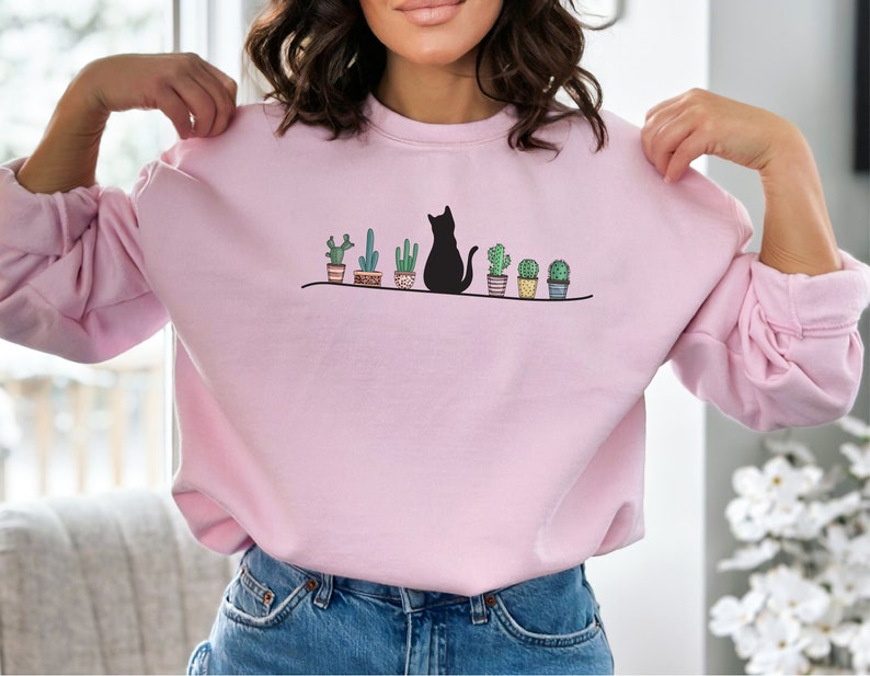 May include: A pink sweatshirt with a black cat sitting in front of a row of potted cacti. The cat is facing left and the cacti are in brown pots with white accents.