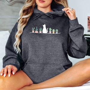 May include: A dark gray hooded sweatshirt with a white cat and potted plants graphic. The cat is sitting with its back to the viewer and looking to the right. The plants are in small pots and have green leaves.