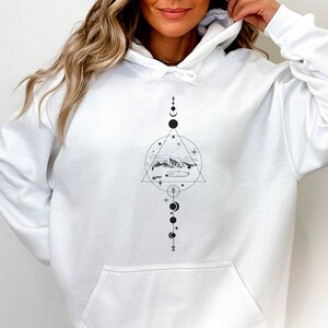 May include: White hoodie featuring a black and white graphic design of a mountain range, clouds, and stars within a triangle and circle. The design also incorporates a vertical line of circles and crescent moons.