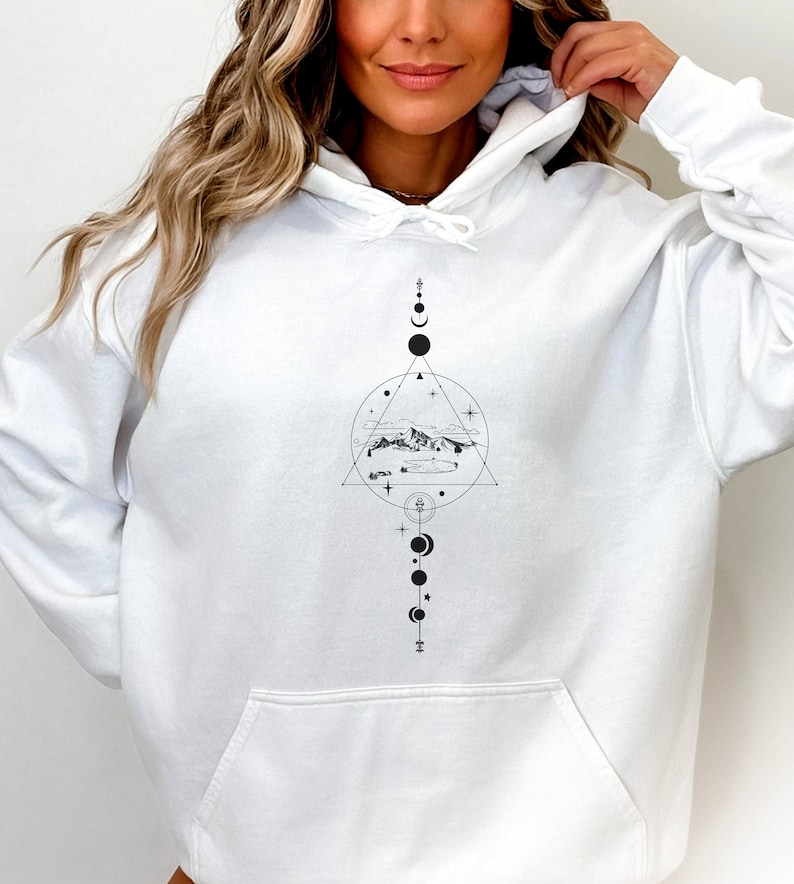 May include: White hoodie featuring a black and white graphic design of a mountain range, clouds, and stars within a triangle and circle. The design also incorporates a vertical line of circles and crescent moons.