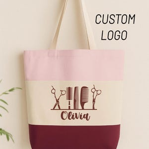 May include: A two-tone tote bag with a light pink top and a burgundy bottom. The center section is off-white with a graphic of scissors, a comb, and a brush. The name "Olivia" is below the graphic, and the text "CUSTOM LOGO" is at the top.
