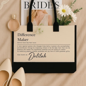 May include: A cream-colored tote bag with black handles and a black bottom panel. The bag features the text "Difference Maker" and "Delilah". A magazine, perfume bottle, and white daisies are also in the image.