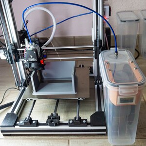 Op de afbeelding: Een 3D-printer met een grijs plastic object dat op het platform wordt geprint. De printer is verbonden met een doorzichtige plastic container met een blauwe slang. De container staat op een houten vloer.