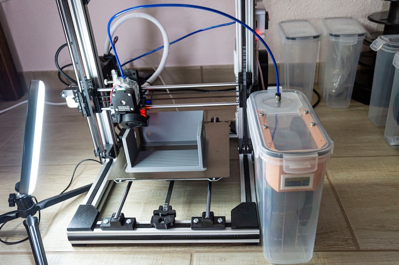 Op de afbeelding: Een 3D-printer met een grijs plastic object dat op het platform wordt geprint. De printer is verbonden met een doorzichtige plastic container met een blauwe slang. De container staat op een houten vloer.