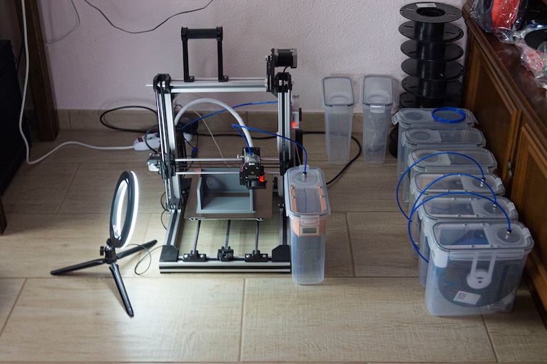 Op de afbeelding: Een 3D-printer met een witte filamentrol, een ringlicht en verschillende doorzichtige plastic containers. De printer staat op een houten vloer.