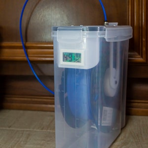 Op de afbeelding: Een doorzichtige plastic container met een blauwe filamentrol erin. De container heeft een digitaal display dat 49 en 18,8% laat zien.