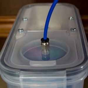 Op de afbeelding: Een doorzichtige plastic container met een blauwe buis die door het deksel loopt. De buis is verbonden met een metalen fitting. De container wordt waarschijnlijk gebruikt voor het opslaan of afgeven van vloeistoffen.