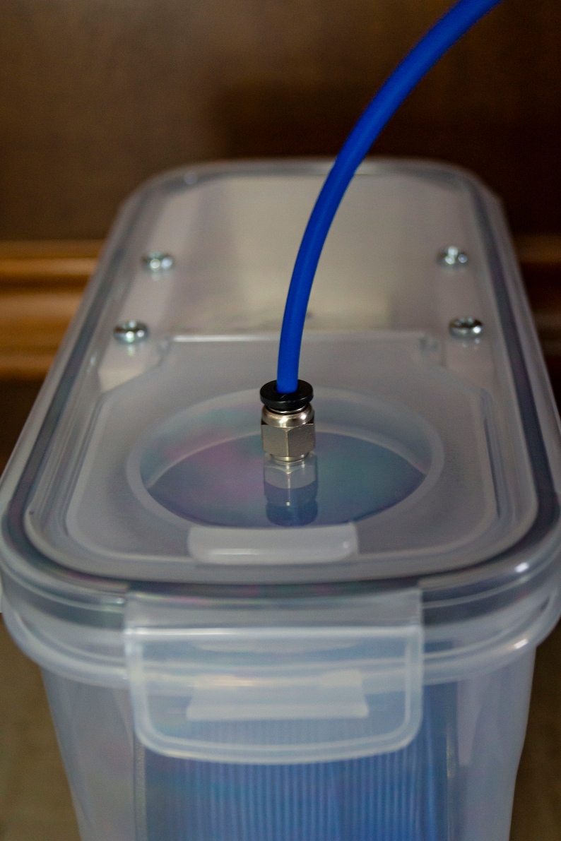 Op de afbeelding: Een doorzichtige plastic container met een blauwe buis die door het deksel loopt. De buis is verbonden met een metalen fitting. De container wordt waarschijnlijk gebruikt voor het opslaan of afgeven van vloeistoffen.