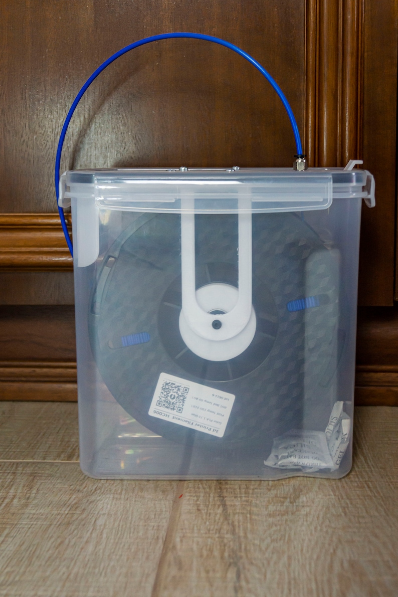 Op de afbeelding: Een doorzichtige plastic container met een blauwe handgreep, die een zwarte spoel filament voor een 3D-printer bevat. De container heeft een label met de tekst "3D Printer Filament, 1,75 mm, PLA, 1 kg".
