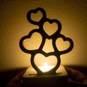 Puede incluir: Un portavelas de metal con forma de árbol con cinco corazones. El portavelas está encendido y la luz brilla a través de los corazones.