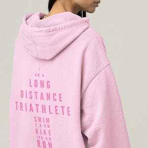 May include: A light pink hoodie with the text "I AM A LONG DISTANCE TRIATHLETE" printed in a darker pink. Below the text are the distances for swim (3.8 km), bike (180 km), and run (42.2 km).