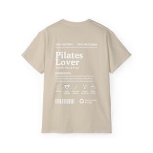 Puede incluir: Camiseta beige con la inscripci&oacute;n blanca "Pilates Lover" y "Studio Flex & Flow". La parte trasera de la camiseta tambi&eacute;n tiene el texto "100% Natural" y "100% Motivated". Texto adicional con "Ingredientes" e iconos.
