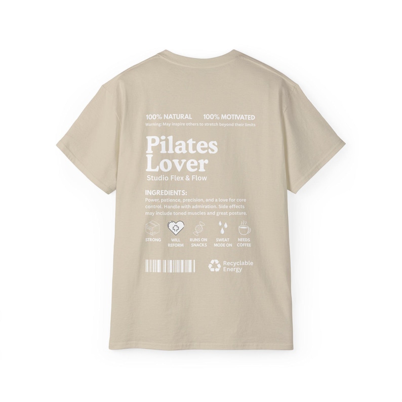 Puede incluir: Camiseta beige con la inscripci&oacute;n blanca "Pilates Lover" y "Studio Flex & Flow". La parte trasera de la camiseta tambi&eacute;n tiene el texto "100% Natural" y "100% Motivated". Texto adicional con "Ingredientes" e iconos.