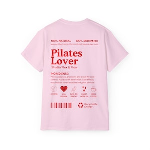 Puede incluir: Una camiseta rosa claro con las palabras "Pilates Lover" en rojo, junto con el texto "Studio Flex & Flow". La camiseta tambi&eacute;n presenta las frases "100% Natural" y "100% Motivado".