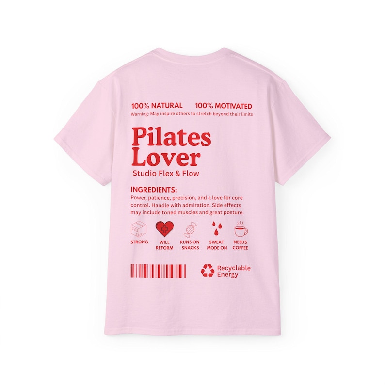 Puede incluir: Una camiseta rosa claro con las palabras "Pilates Lover" en rojo, junto con el texto "Studio Flex & Flow". La camiseta tambi&eacute;n presenta las frases "100% Natural" y "100% Motivado".