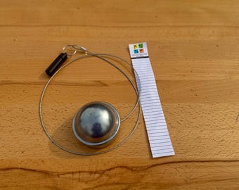 May include: A silver-colored metal sink stopper with a black handle, attached to a wire cable. A small notepad with a colorful logo is also present. The items are on a wooden surface.