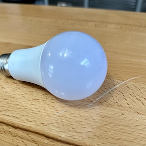 May include: A white LED light bulb with a rounded, frosted glass bulb and a silver base. The light bulb is resting on a wooden surface.
