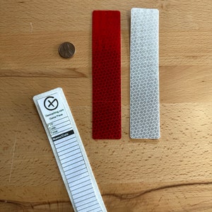 May include: Three rectangular geocaching game pieces and a copper penny. One piece is white with a black Geocaching.com logo and lines for FTF, Date, Time, and Name/Date. The other two pieces are solid red and silver with a hexagonal grid pattern.