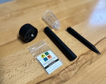 May include: A black and clear plastic geocaching log book container with a black metal stake, a black plastic cap, and a clear plastic lid. The log book is labeled "Official Geocaching Log Book" with the words "Date/Name" printed below.