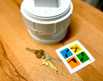 May include: A white PVC pipe cleanout plug with a square nut top, a set of two brass-colored metal keys on a ring, and a square white sticker featuring a four-quadrant design with yellow, blue, green, and orange sections and a black graphic.