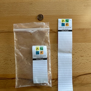 May include: Two official geocaching log books, one in a clear plastic bag and the other on a wooden surface. Both log books have a colorful logo with a person walking, a compass, and a flag.