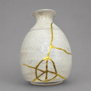 May include: A small, off-white ceramic vase with a speckled texture and a narrow neck. The surface features prominent, jagged cracks repaired with a bright, reflective gold-colored lacquer, creating a distinct web-like pattern across the body.