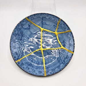 May include: A round blue ceramic plate featuring a raised relief landscape design of mountains and trees. The plate is repaired with prominent gold-colored lines that crisscross the surface, highlighting the fractures in the ceramic material.