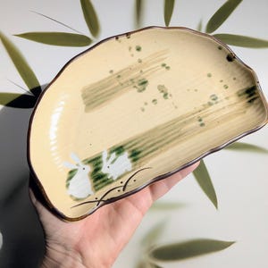 May include: Cream-colored ceramic serving dish with a brown rim and an irregular shape. The dish features a painted design of two white rabbits and green brushstrokes. The background includes bamboo leaves.