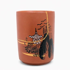 May include: A terracotta-colored ceramic cup with a hand-painted Japanese landscape design. The artwork features a pagoda, mountains, and flying birds, with gold accents and visible kintsugi repair lines.