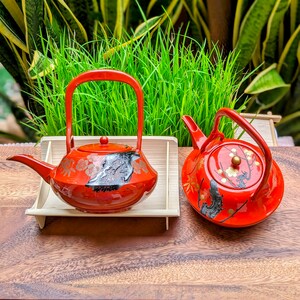 Op de afbeelding: Twee rode Japanse theepotten met bijpassende rode handvatten. Eén theepot staat op een bamboe dienblad. De theepotten hebben bloemen en abstracte ontwerpen in goud en zwart. De theepotten staan op een houten oppervlak.