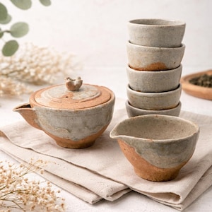 May include: A ceramic tea set with a teapot, a serving bowl, and a stack of small teacups. The set has a rustic, handmade appearance with a mix of tan, gray, and brown tones. The teapot has a lid and a spout. The teacups are stacked.