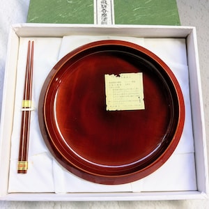 以下が含まれることがあります： 丸い赤褐色の漆塗りのトレイと箸のセットが入った箱。箸は茶色で金色のアクセントが付いています。箱には、日本語のテキストが入った緑色の蓋と白い内装が含まれています。