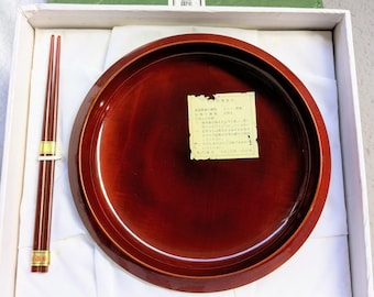 May include: A boxed set featuring a round, reddish-brown lacquered tray and a pair of chopsticks. The chopsticks are brown with gold accents. The box includes a green lid with Japanese text and a white interior.