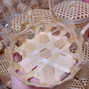 Puede incluir: Una cesta de bambú tejida a mano, de color marrón claro, con un diseño geométrico único. La cesta redonda tiene la parte superior abierta y se sostiene en una mano. Otras cestas similares son visibles en el fondo, lo que sugiere una colección o una exhibición de mercado.