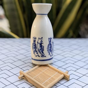 May include: A white ceramic sake bottle with blue fish illustrations and a blue rim, paired with a small square bamboo coaster. The bottle has a unique shape, and the coaster adds a natural touch.