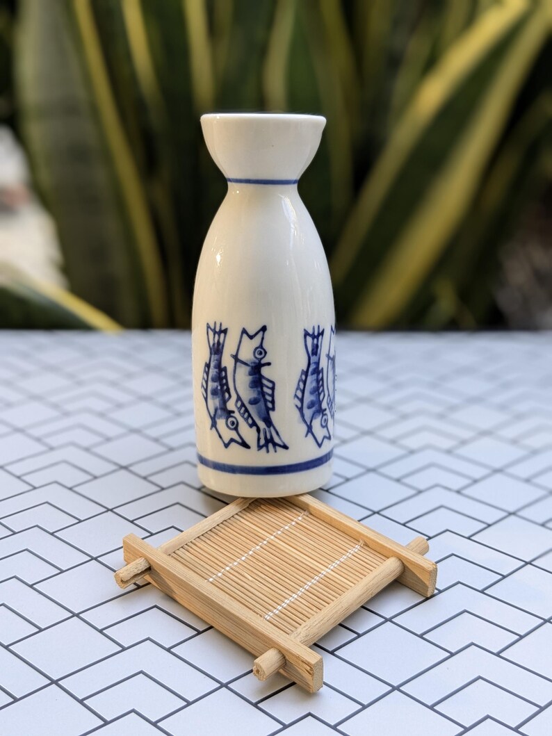 May include: A white ceramic sake bottle with blue fish illustrations and a blue rim, paired with a small square bamboo coaster. The bottle has a unique shape, and the coaster adds a natural touch.