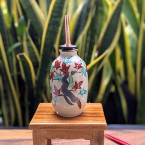 May include: A ceramic oil bottle with a copper spout, decorated with a tree and red, blue, and green leaves. The bottle sits on a small wooden table, with a blurred green background.