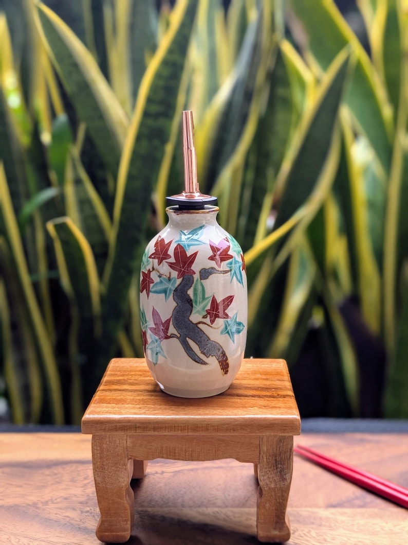 May include: A ceramic oil bottle with a copper spout, decorated with a tree and red, blue, and green leaves. The bottle sits on a small wooden table, with a blurred green background.