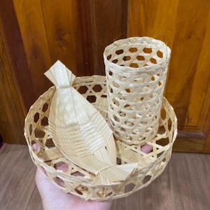May include: A collection of three bamboo woven items: a round basket, a cylindrical cup, and a fish-shaped ornament. The basket and cup feature an open, lattice-like pattern. The items are light brown in colour.