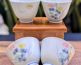 Könnte beinhalten: Set aus vier weißen Keramik-Teetassen mit blauen und floralen Designs. Die Tassen haben einen blauen Rand und ein kleines goldenes Etikett mit der Aufschrift "有田焼 ARITA".