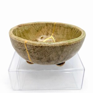 Op de afbeelding: Een kleine, ronde keramische kom met een getextureerde, aarde-bruine buitenkant en een groene binnenkant. De kom heeft drie kleine voetjes en is gerepareerd met goudkleurige kintsugi. De kom wordt gepresenteerd op een heldere acryl standaard.