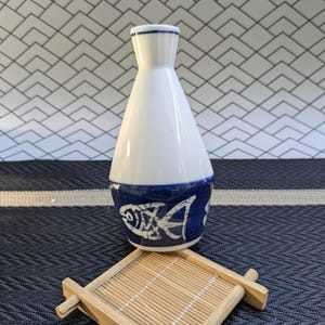 May include: A white ceramic sake carafe with a blue fish design around the base. The carafe sits on a small, square bamboo coaster. The background features a geometric tile pattern.