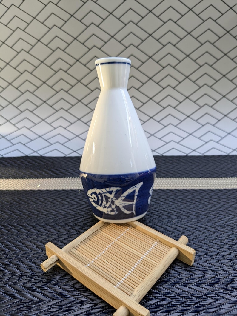 May include: A white ceramic sake carafe with a blue fish design around the base. The carafe sits on a small, square bamboo coaster. The background features a geometric tile pattern.