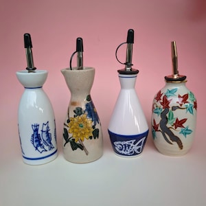 Puede incluir: Cuatro botellas de cerámica para aceite y vinagre con boquillas de acero inoxidable. Las botellas presentan varios diseños, incluyendo peces azules, flores amarillas y un árbol con hojas rojas. Sobre fondo rosa.