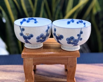 Könnte beinhalten: Zwei kleine weiße Keramikschalen mit blauen und goldenen Blumendesigns. Die Schalen stehen auf einem kleinen Holzständer.