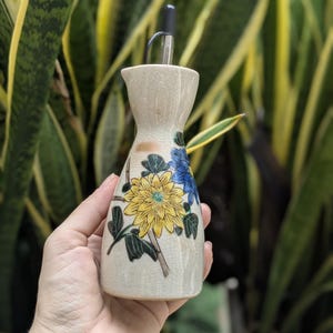 May include: A beige ceramic oil bottle with a black spout. The bottle is decorated with yellow and blue floral designs and green leaves. The bottle is held in a hand against a background of green and yellow foliage.