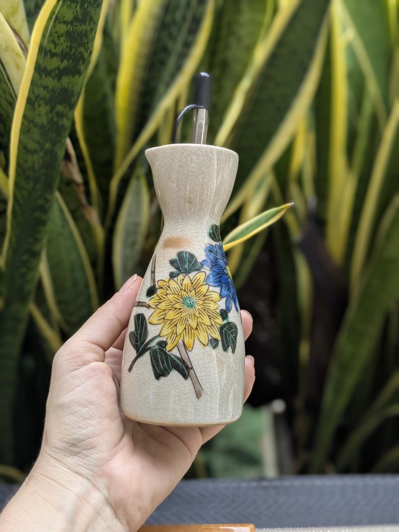 May include: A beige ceramic oil bottle with a black spout. The bottle is decorated with yellow and blue floral designs and green leaves. The bottle is held in a hand against a background of green and yellow foliage.