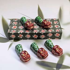 Puede incluir: Juego de seis reposa palillos de cerámica. Cada reposa palillos presenta una cabeza de dragón roja con detalles dorados y un cuerpo verde estampado. Los reposa palillos se muestran con una caja estampada.