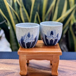 May include: Two ceramic cups with a speckled, light blue and white finish, each adorned with a blue floral design. The cups sit on a small, wooden table with curved legs, showcasing a handcrafted aesthetic. The background features lush green foliage.